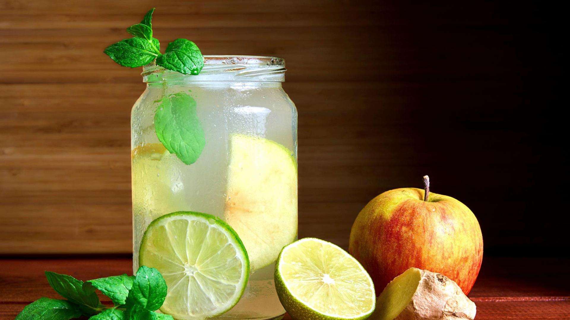 kalter hausgemachter Switchel-Drink in einem Marmeladenglas mit Limetten, Ingwer & Apfel neben dem Glas