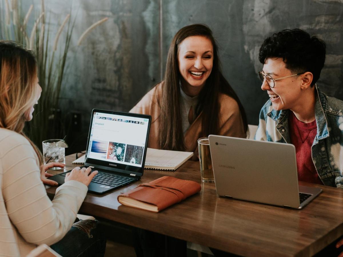 Drei Frauen sitzen lachend um einem Tisch mit ihren Laptops