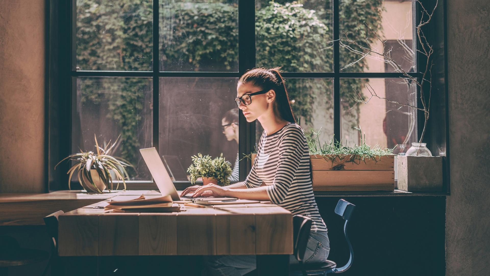 junge Frau mit Brille sitzt an vor einem Laptop am Fenster in einem Büro oder Café