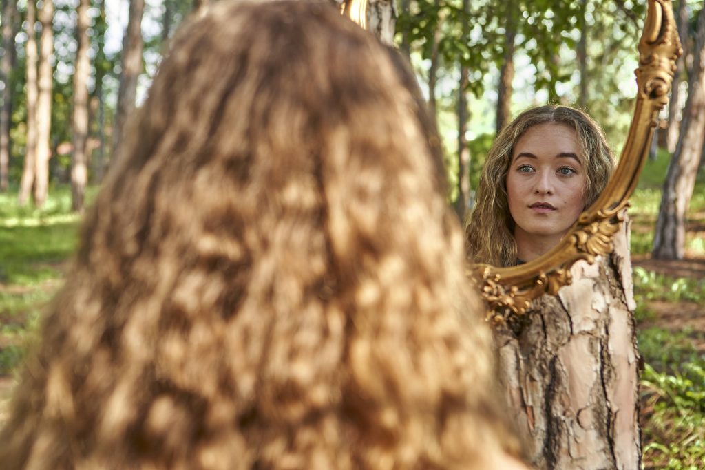 Blonde Frau steht im Wald vor einem Spiegel