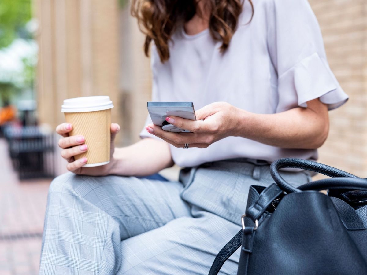 Frau sitzt mit Kaffee und Handy in der Hand auf einer Bank
