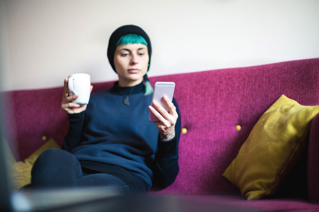Junge Frau sitzt auf Couch und schaut auf Smartphone