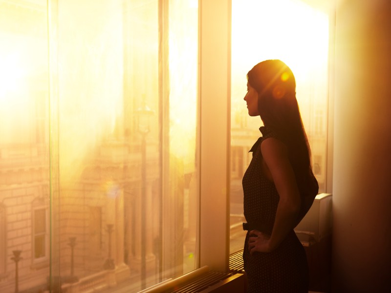 Eine Frau steht im Büro vor dem Fenster. Es ist Sonnenuntergangsstimmung.