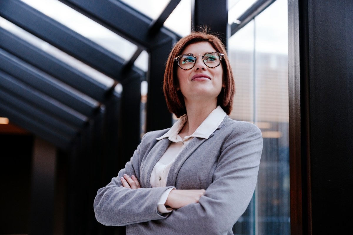 Eine Business-Frau steht mit verschränkten Armen vor einem Fenster.