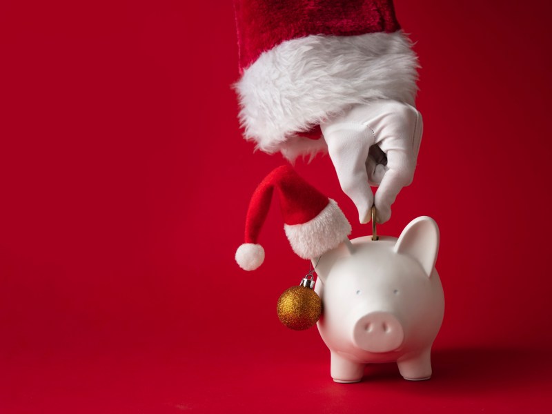 Eine Hand schmeißt Geld in ein Sparschwein mit Weihnachtsmütze.