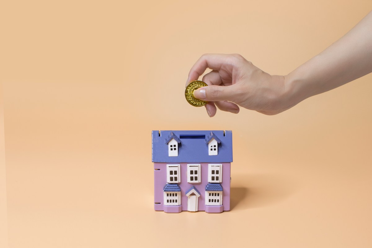 Hand wirft Münze in ein Sparschwein in Form eines Hauses.