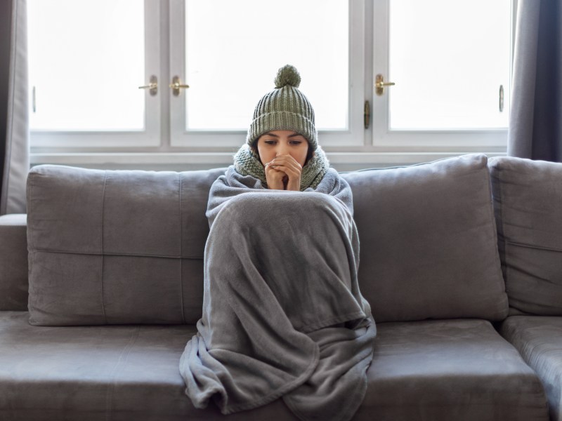 Eine Frau sitzt mit einer decke auf dem Sofa. Sie trägt eine Münze und friert.