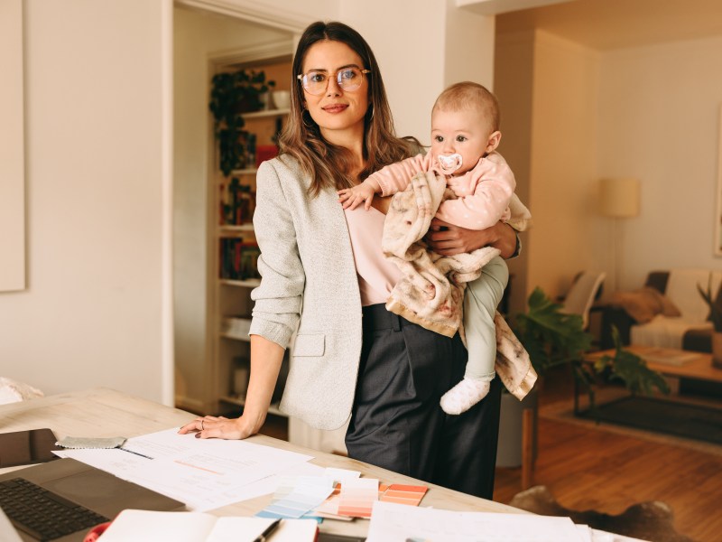 Eine Frau hält ein Baby auf dem Arm und steht vor einem Schreibtisch.