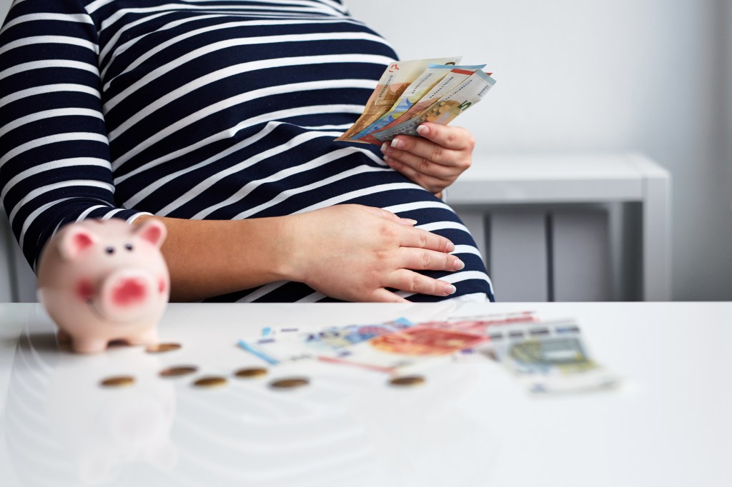 Eine Schwangere sitzt an einem Tisch. Auf diesem liegt Geld.
