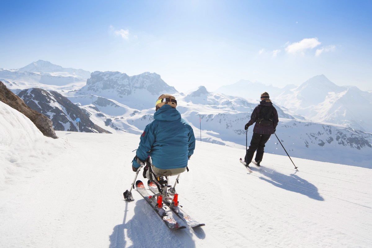 Zwei Skifahrer fahren die Piste runter. Im Hintergrund sind Berge zu sehen.
