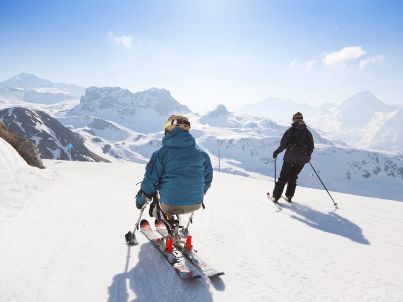 Zwei Skifahrer fahren die Piste runter. Im Hintergrund sind Berge zu sehen.