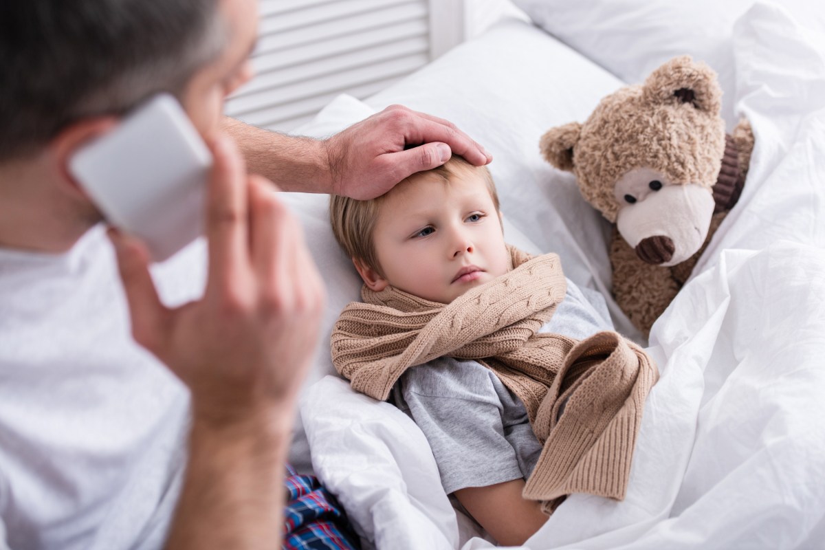 Ein Kind liegt krank im Bett. Der Vater hält die Hand auf seine Stirn.