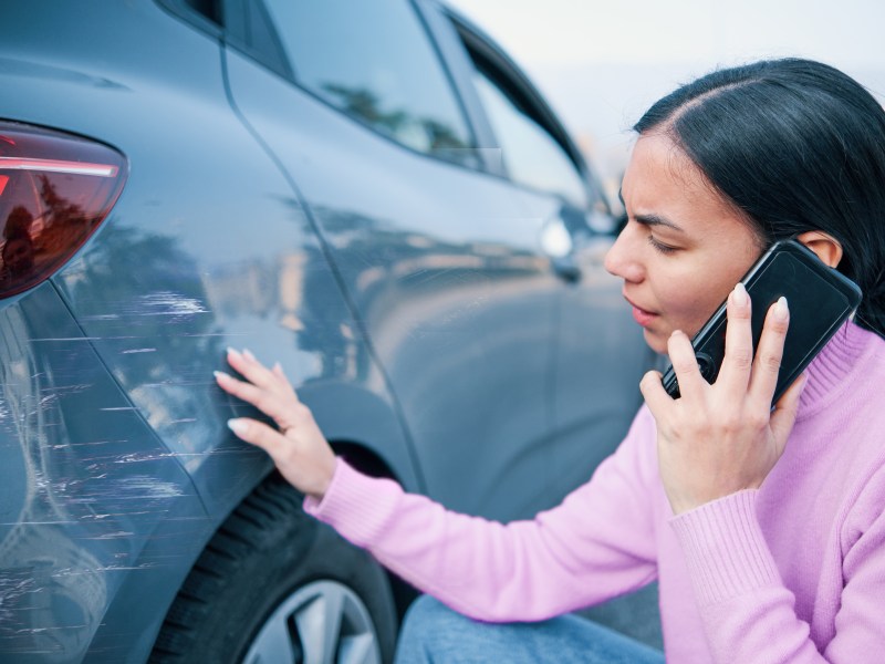 Frau telefoniert und schaut sich ihren Schaden am Auto an.