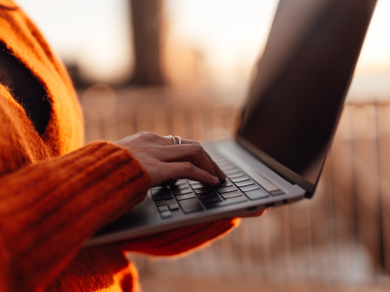 Frau tippt mit der Hand auf einer Laptop-Tastatur in einer Außenumgebung in der Stadt.