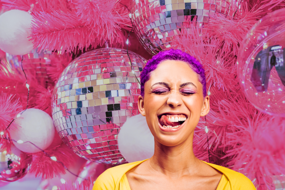 Eine Frau mit kurzen, lila gefärbten Haaren zieht eine Grimasse. Sie steht vor einem pinken Weihnachtsbaum mit Disco-Kugeln.