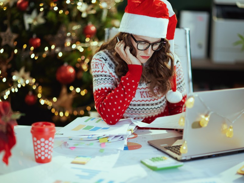 Frau sitzt vor Laptop. Die Stimmung ist weihnachtlich. Hinten steht ein Weihnachtsbaum.