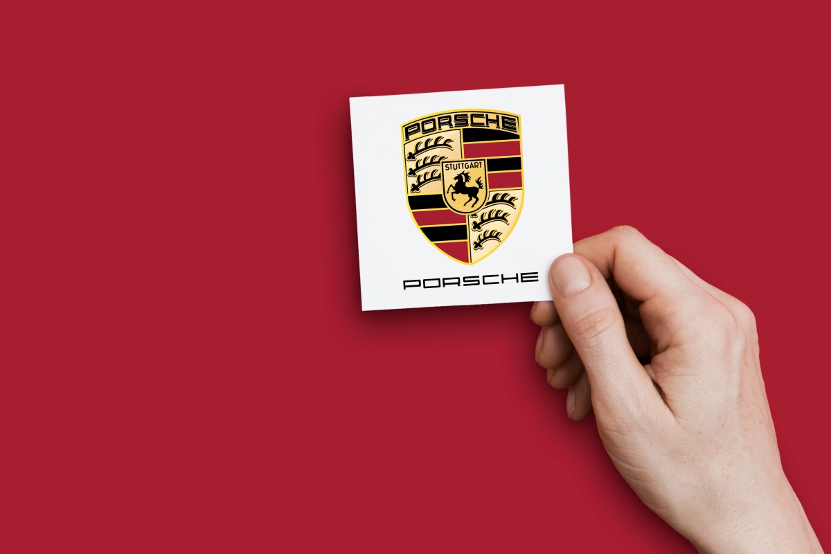 Eine Hand hält das Logo von Porsche vor eine rote Wand.