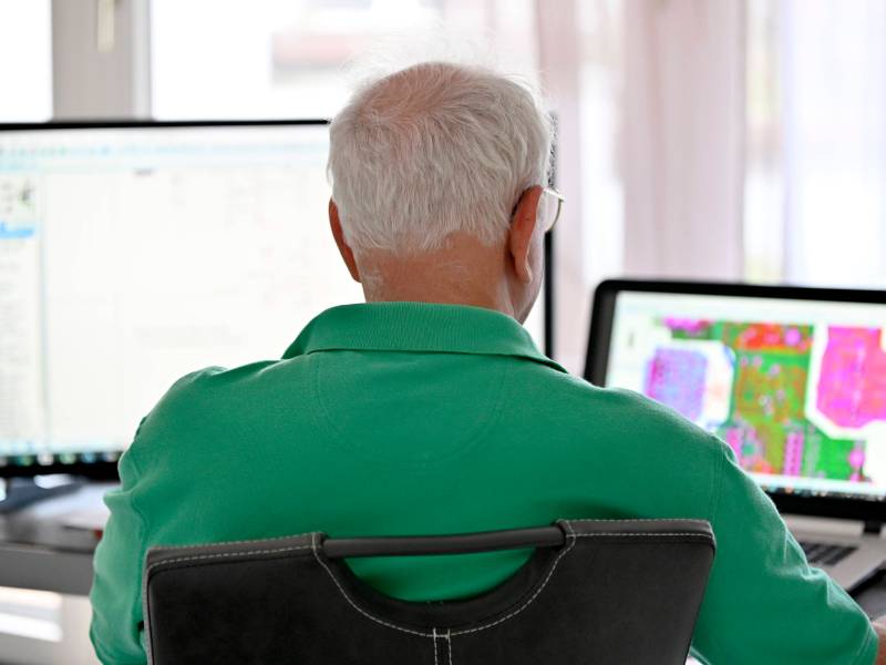 Ein älterer Mann sitzt vor zwei Computerbildschirmen.