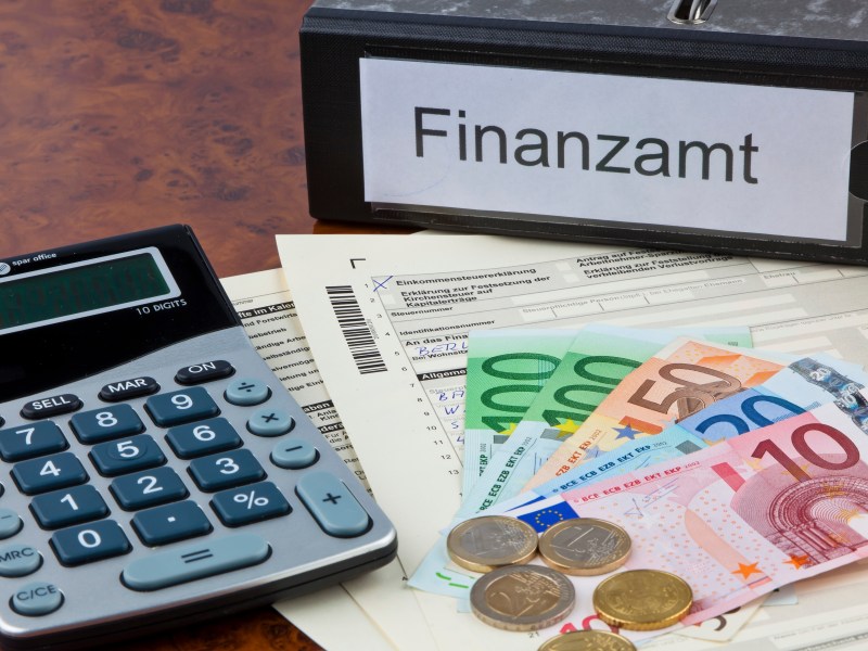 Ein Taschenrechner, Geld und Steuerunterlagen liegen neben einem Ordner mit der Aufschrift "Finanzamt".