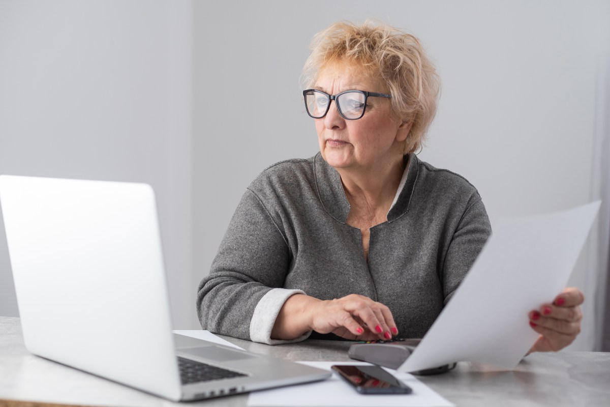 Eine Seniorin sitzt mit Dokumenten am Laptop. Sie sieht konzentriert aus.