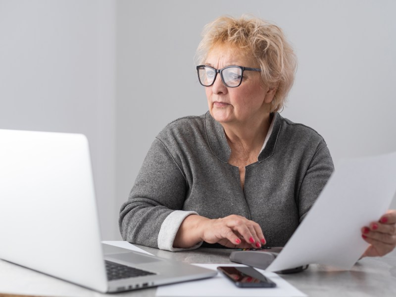 Eine Seniorin sitzt mit Dokumenten am Laptop. Sie sieht konzentriert aus.