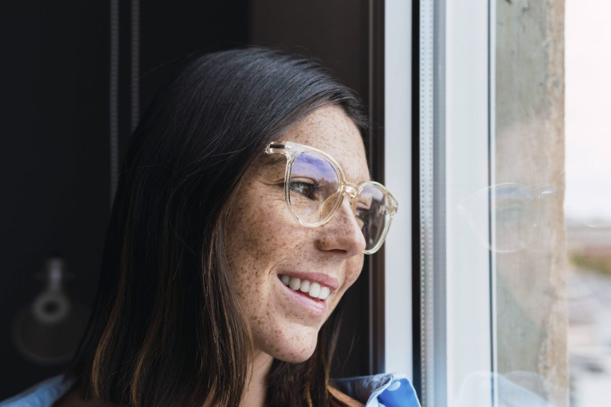 Eine Frau mit Sommersprossen und Brille blickt aus dem Fenster.