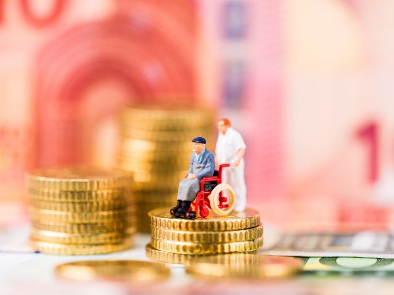 Eine Spielfigur, Mann im Rollstuhl und Pfleger, stehen auf einem Münzhaufen.