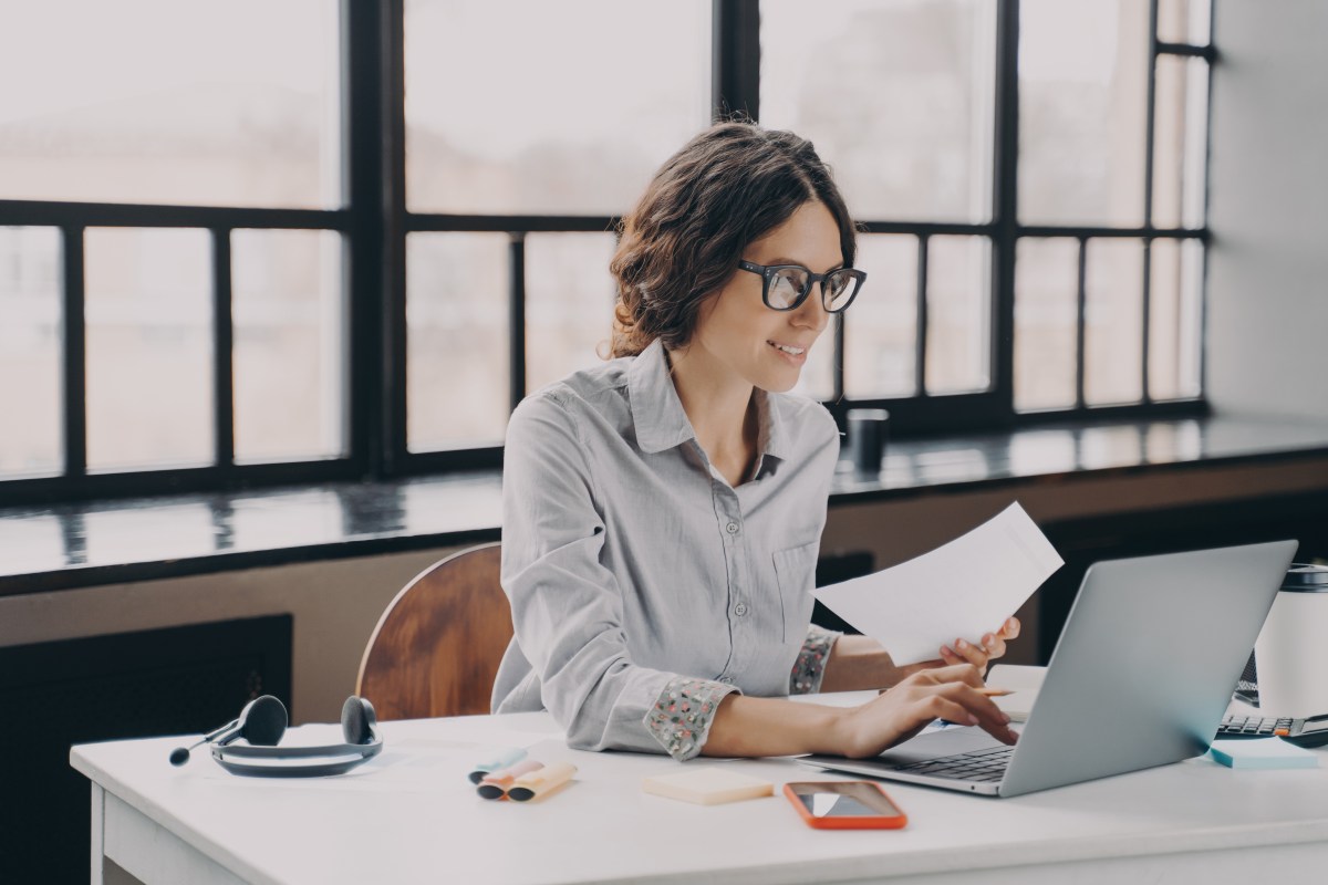 Eine Frau sitzt am Laptop und hält ein Dokument in der Hand.