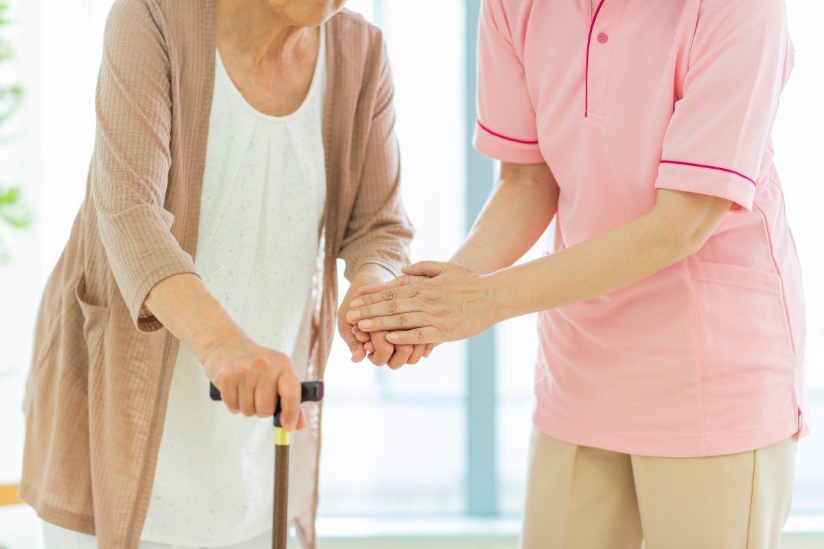 Eine Pflegerin in einem rosafarbenen Kittel hilft einer älteren Dame mit Gehstock.
