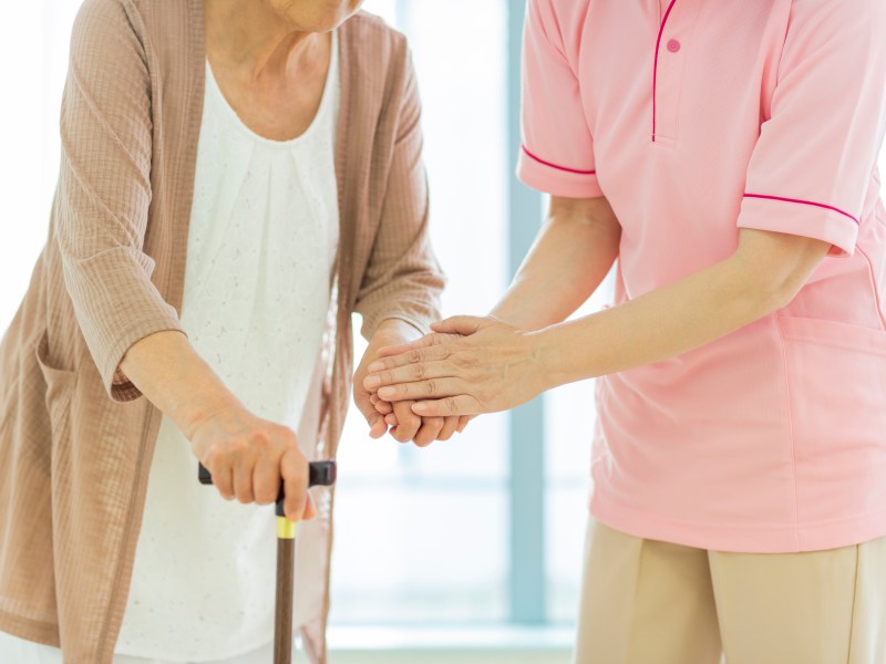 Eine Pflegerin in einem rosafarbenen Kittel hilft einer älteren Dame mit Gehstock.