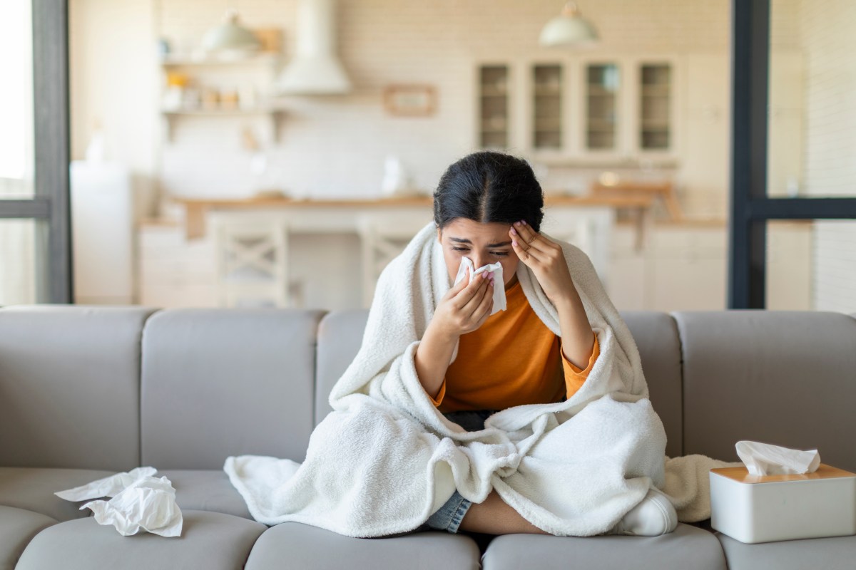 Eine Frau sitzt erkältet auf dem Sofa. Sie putzt sich die Nase.