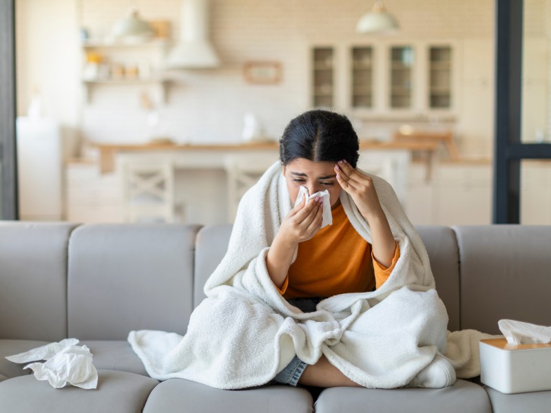 Eine Frau sitzt erkältet auf dem Sofa. Sie putzt sich die Nase.