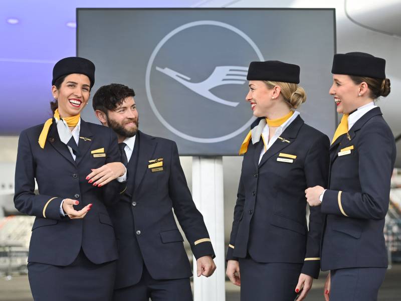 Eine Gruppe von Flugbegleiterinnen und einem Flugbegleiter stehen vor dem Logo der Lufthansa.