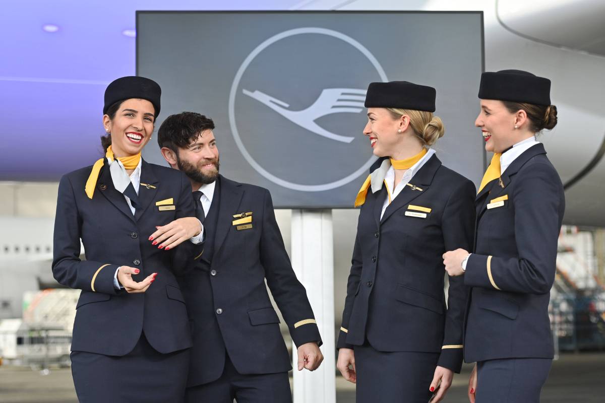 Eine Gruppe von Flugbegleiterinnen und einem Flugbegleiter stehen vor dem Logo der Lufthansa.