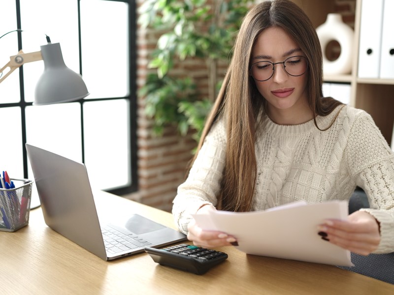 Eine Frau sitzt am Schreibtisch und schaut sich ein Dokument an. Neben ihr steht ein Laptop.