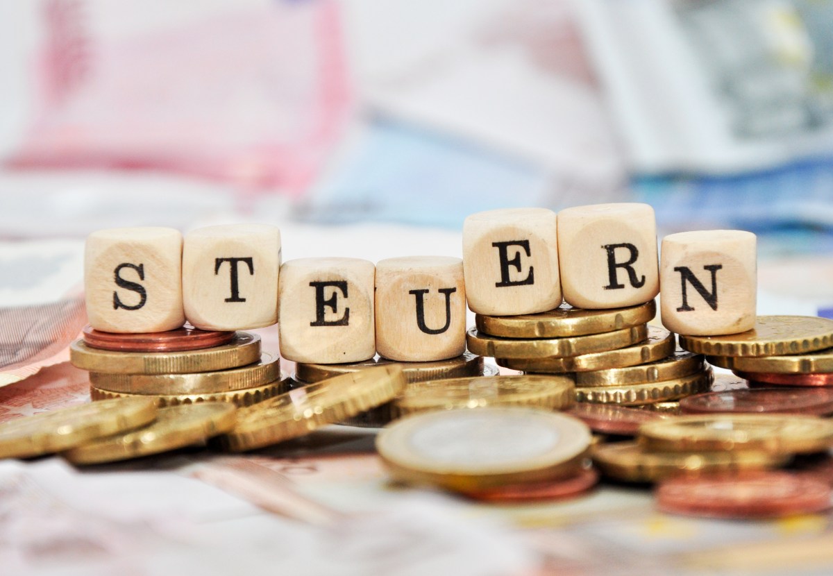 Mit Buchstaben aus Holzwürfeln würde das Wort Steuer geschrieben. Sie stehen auf einem Stapel Münzen.