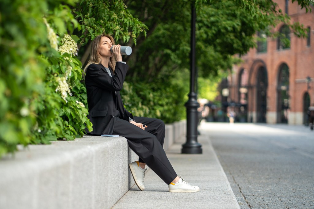 Eine Frau sitzt auf einer Mauer im Park und trinkt aus einer Flasche.