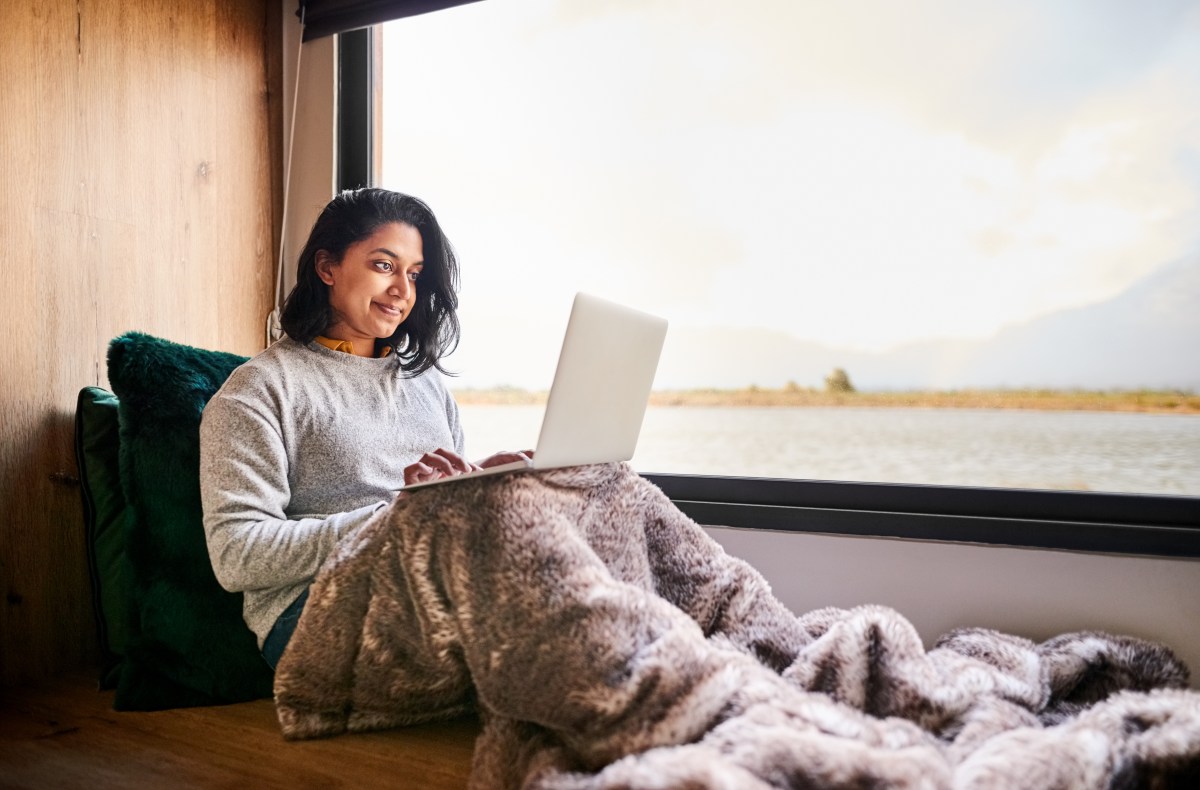 Frau sitzt mit Decke am großen Fenster und benutzt Laptop zu Hause.