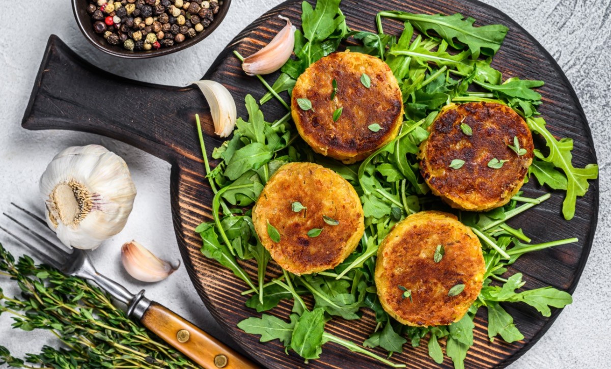 Vier Linsenfrikadellen auf einem Bett aus Rucola auf einem runden Holzbrett. Daneben steht eine Schale mit Pfeffer und es liegen Knoblauchzehen herum.