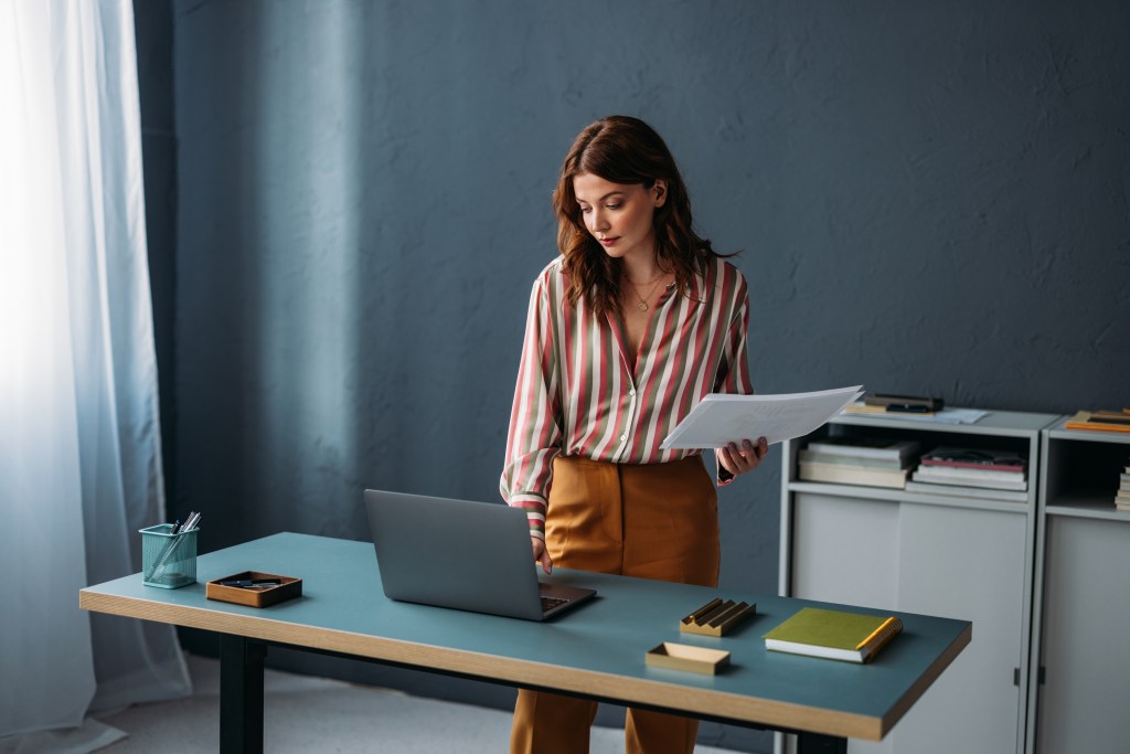 Konzentrierte, berufstätige Frau steht mit Laptop und Papierkram an einem Schreibtisch in einem modernen Büroarbeitsplatz.