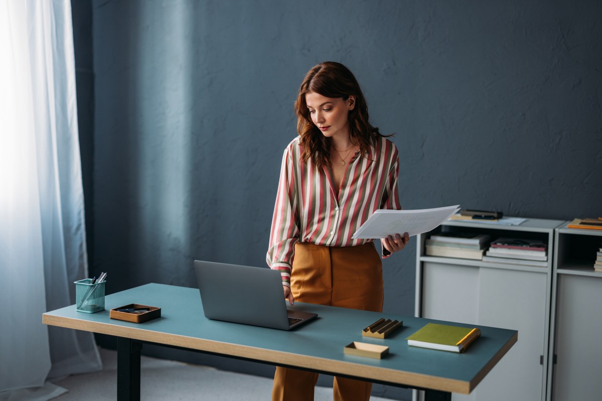 Konzentrierte, berufstätige Frau steht mit Laptop und Papierkram an einem Schreibtisch in einem modernen Büroarbeitsplatz.