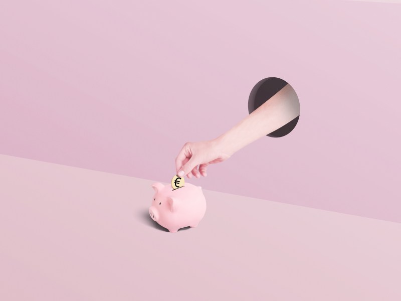 Hand legt eine goldene Euro-Münze in ein rosa Sparschwein.