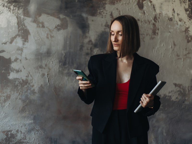 Dreiviertelaufnahme einer Geschäftsfrau in einer schwarzen Jacke, die ihr Smartphone benutzt und ein Tablet in der Hand hält.