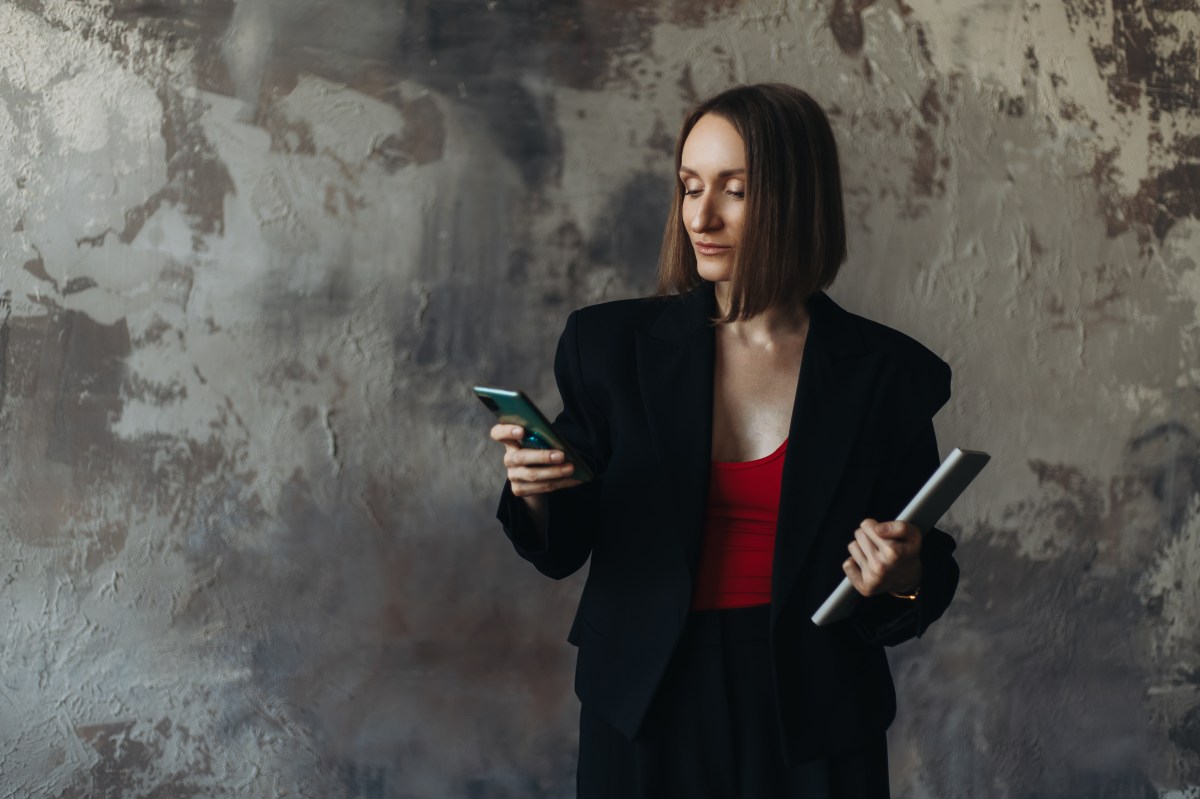 Dreiviertelaufnahme einer Geschäftsfrau in einer schwarzen Jacke, die ihr Smartphone benutzt und ein Tablet in der Hand hält.