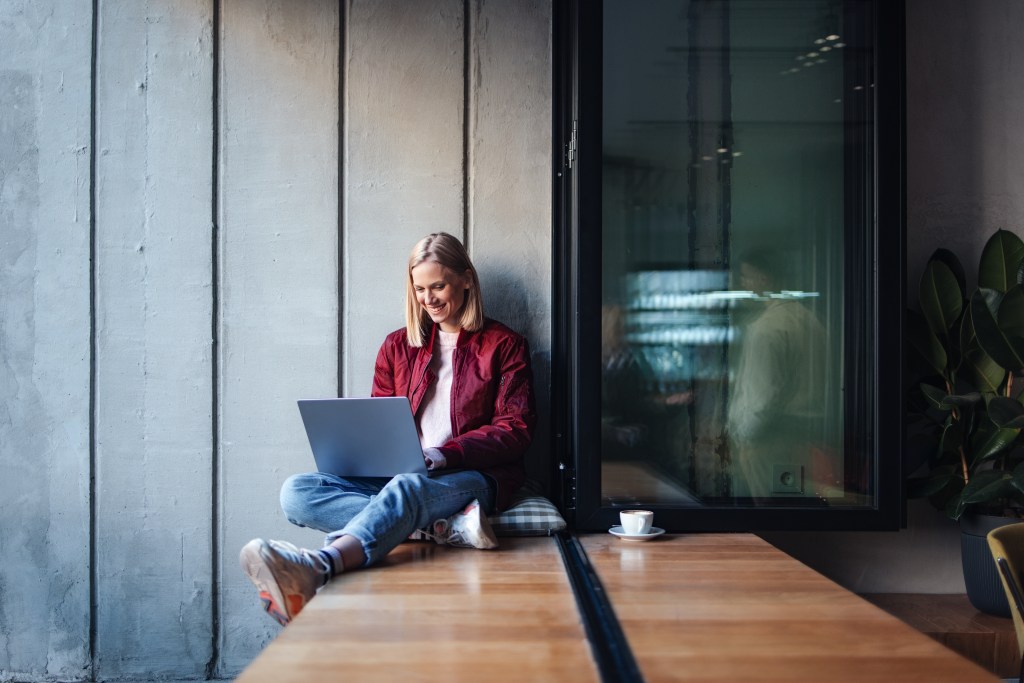Eine junge Frau in einem lässigen Outfit lächelt, während sie ihren Laptop an einem gemütlichen Fensterplatz im Café benutzt.