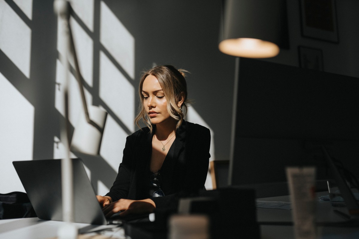 Eine junge Frau mit blonden Haaren sitzt im BÃ¼ro und arbeitet am Laptop.