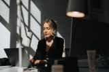 Eine junge Frau mit blonden Haaren sitzt im BÃ¼ro und arbeitet am Laptop.