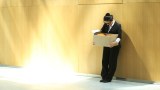 Eine Frau im Anzug lehnt an der Wand und schaut zu Boden. Sie trägt einen Karton mit Sachen.