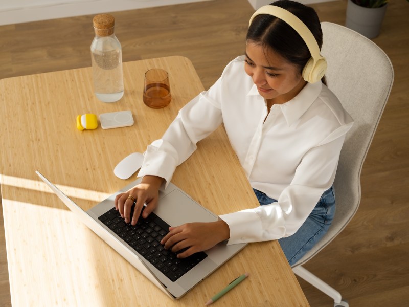 Junge Frau mit Kopfhörern arbeitet zu Hause mit Laptop am Tisch.