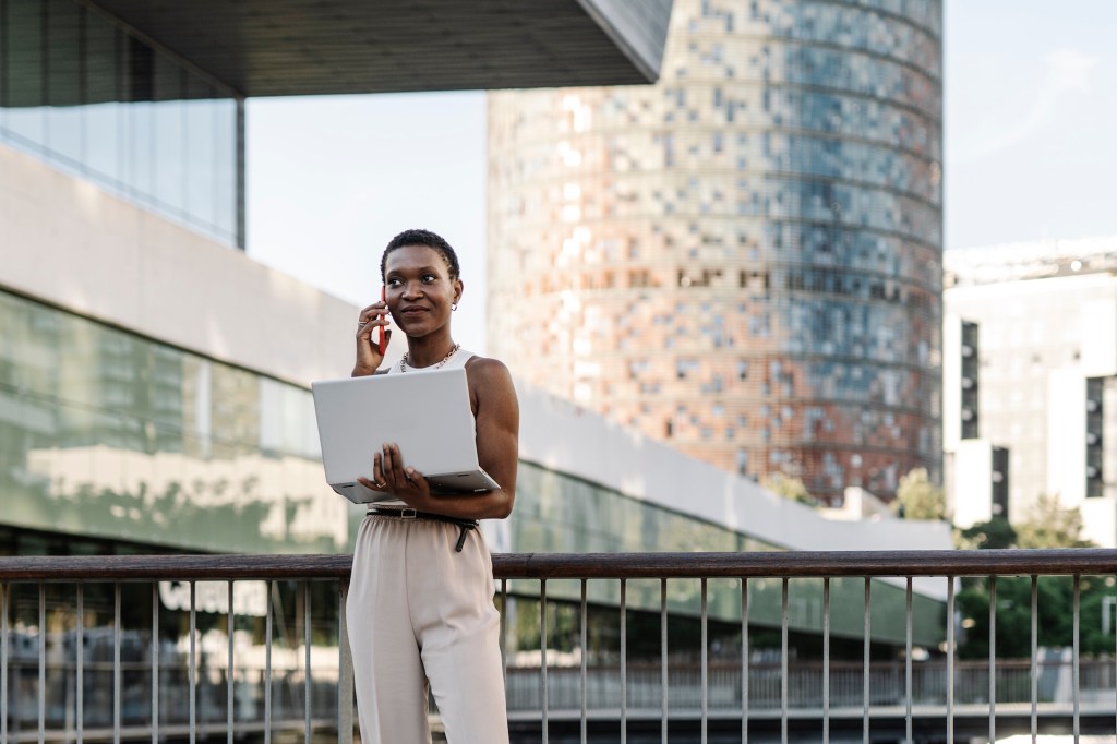 Frau mit Laptop, die am Geländer steht und mit dem Handy telefoniert.